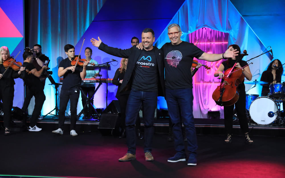 Andrew Bell, CPO of Kinaxis and John Sicard, CEO stand on stage at Kinexions 24 with an orchestra behind them.