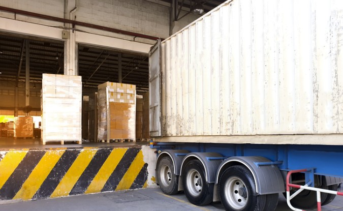 Stack of cardboard boxes wrapped in plastic sitting on a loading dock next to a semi truck at a distribution warehouse.