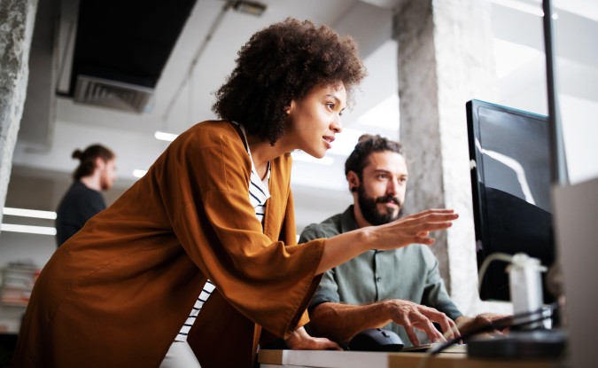 Woman in gold jacket leans toward computer monitor, with a bearded man seated to her right.