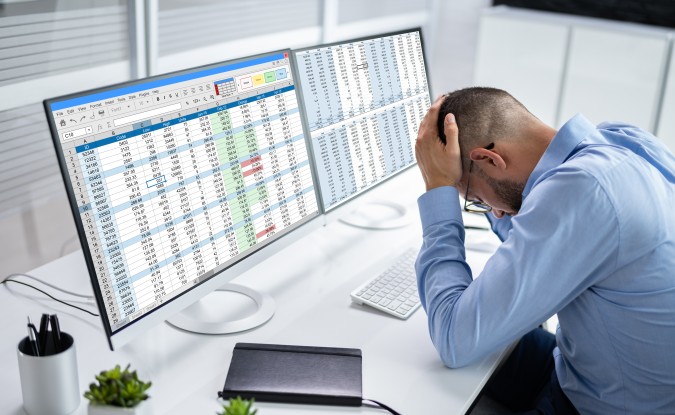 Man sits at a desk in front of two computer screens displaying spreadsheets. He is holding his head in a manner that indicates stress or defeat.