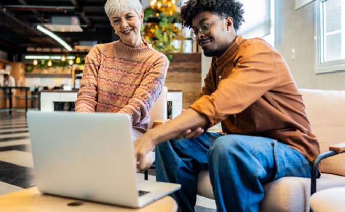 An older white woman sits next to a younger Black man, both of them dressed casually in an office lobby. They are smiling together at a laptop screen. 