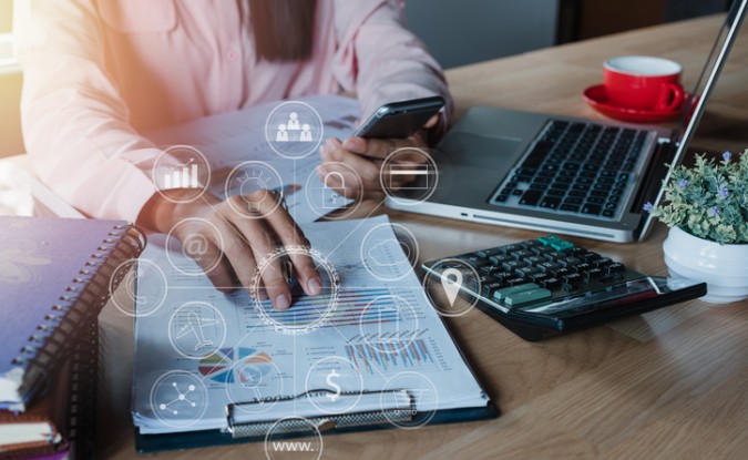 A businesswoman working with her smartphone, laptop and calculator in an office. Financial data and iconography are overlaid on the photo.