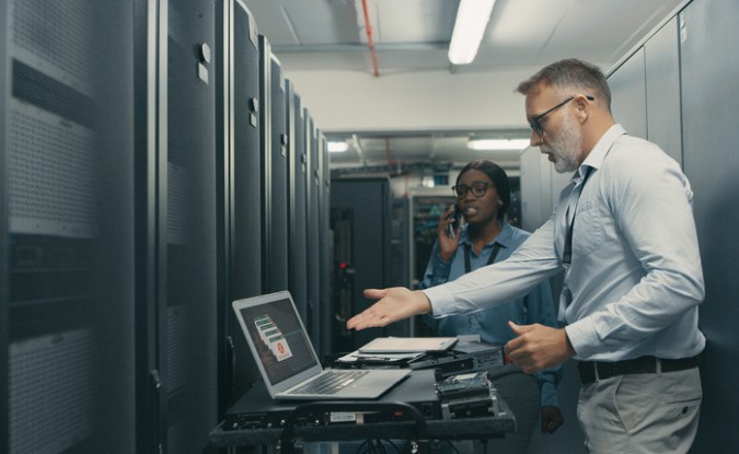 A man and a woman standing in a server room. The man gestures frustratedly to his laptop screen, which displays multiple error messages. The woman is speaking into a cell phone. 